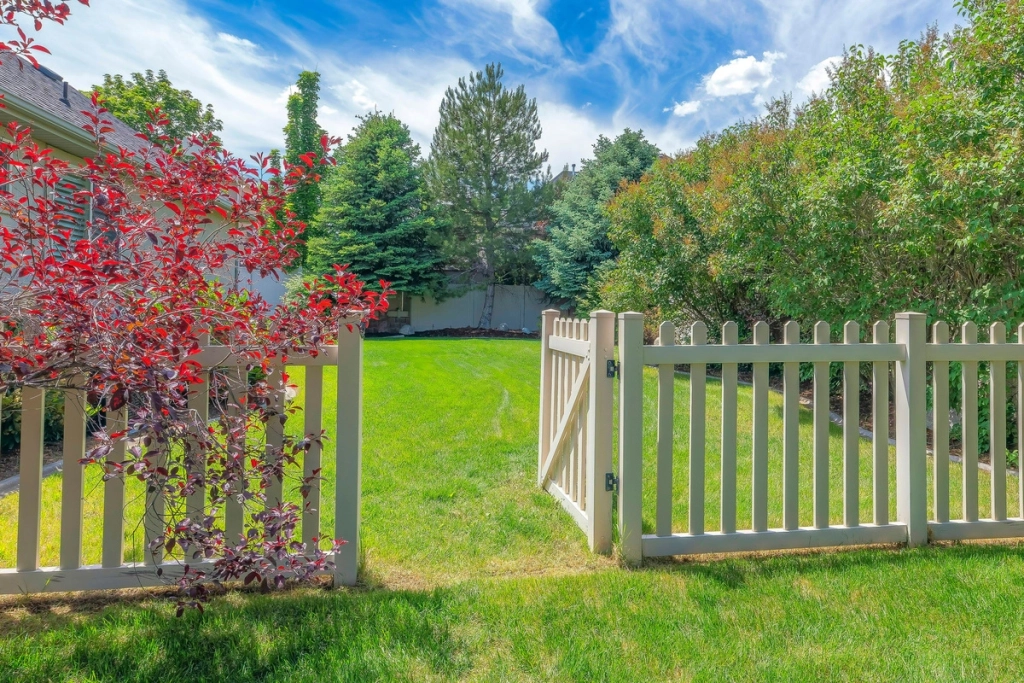 Clôture et portail en bois clair entourant un jardin verdoyant avec des arbres et des arbustes, offrant sécurité et esthétique à la propriété.