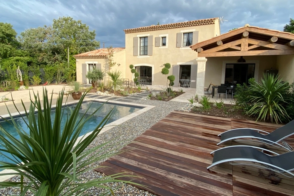 Aménagement extérieur avec piscine, terrasse en bois et jardin paysager devant une maison de style méditerranéen.